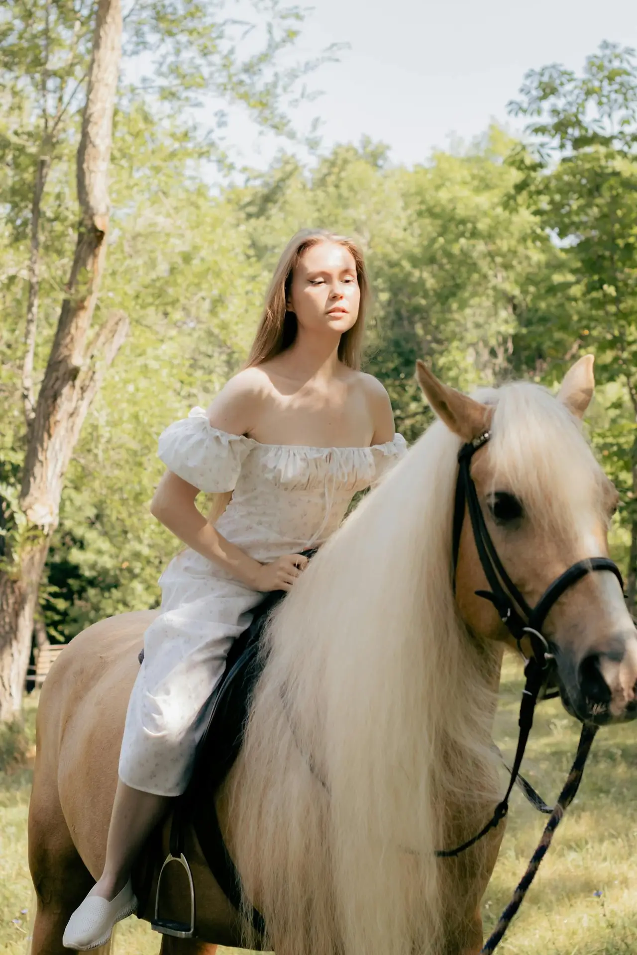 A young woman in an off-shoulder dress rides a palomino horse in a sunny rural setting.
